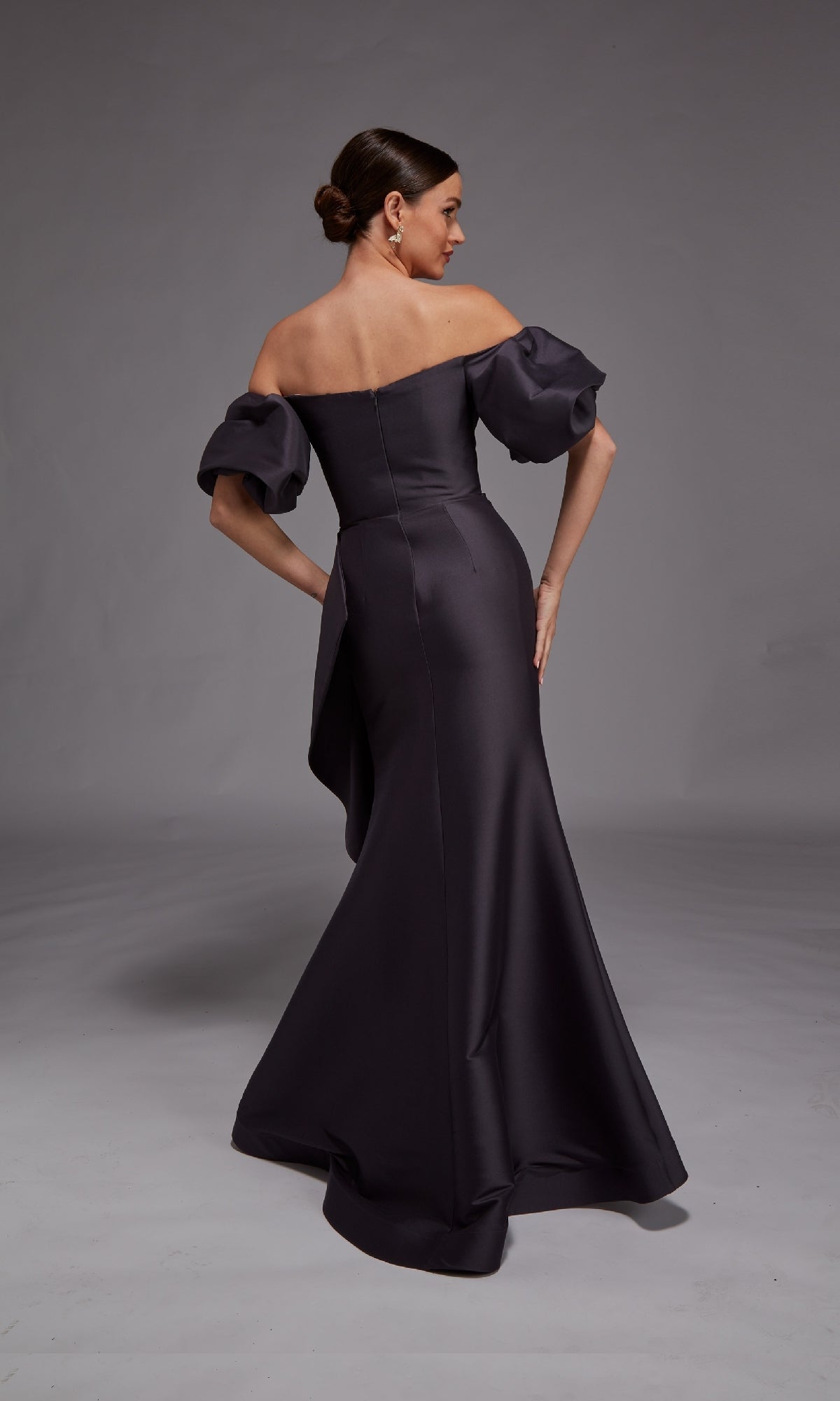 A woman stands facing away against a neutral backdrop, modeling the Alyce 27863 long formal dress—an off-the-shoulder design with puffed sleeves and a fitted silhouette.